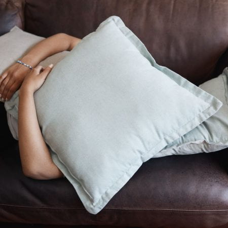 Tired, depressed girl sleeping under pillow on sofa in the living room. Woman hide feeling sad or anxiety and problem with depression, mental health and fatigue or fear on couch in the lounge